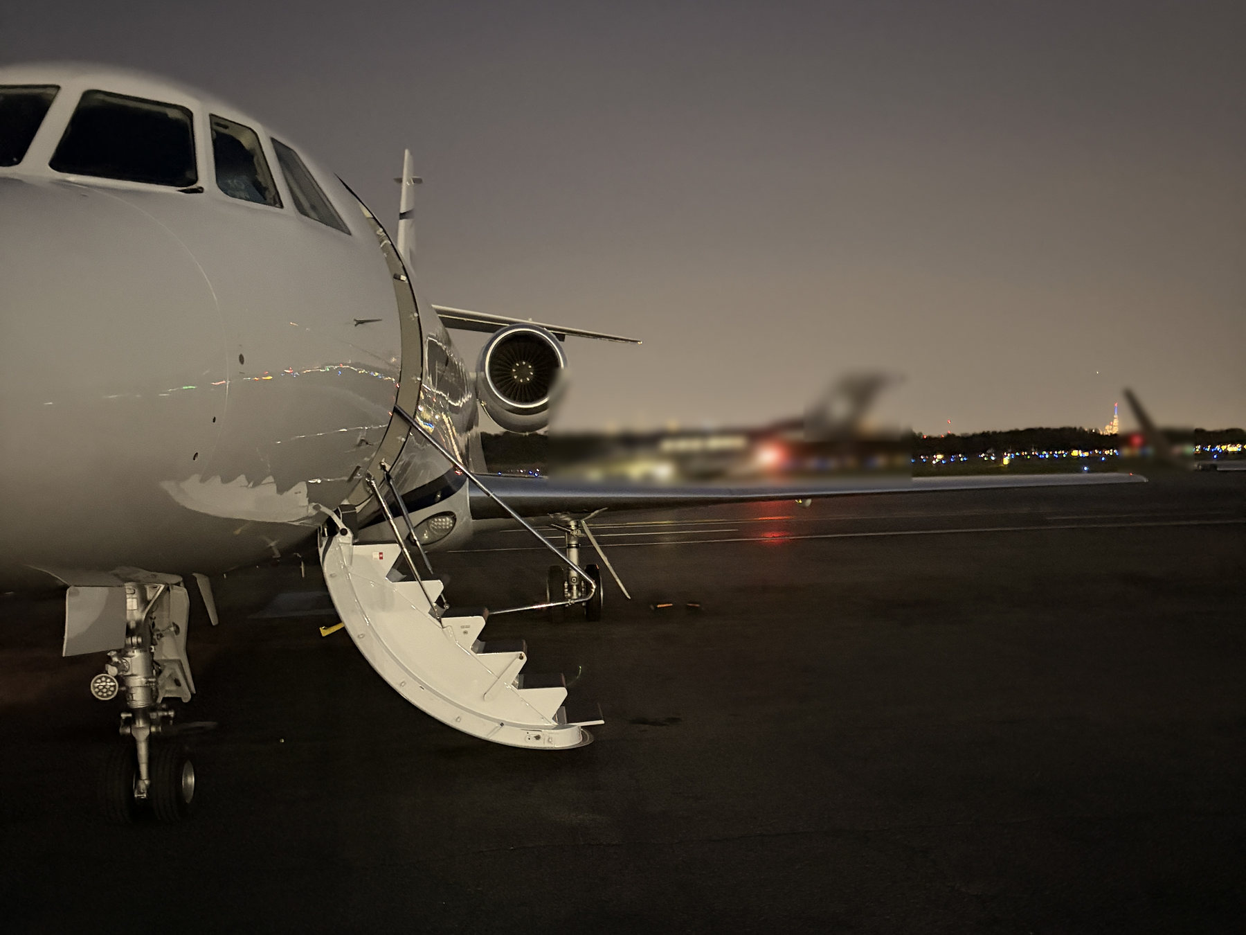 Falcon business jet with the airstair down at dusk, other jets and a city skyline behind